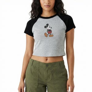 Op de afbeelding: Een grijze crop top met zwarte korte mouwen met een Mickey Mouse-afbeelding. Het stripfiguur is afgebeeld in zijn iconische rode shorts, gele schoenen en witte handschoenen. Het shirt heeft een casual stijl.