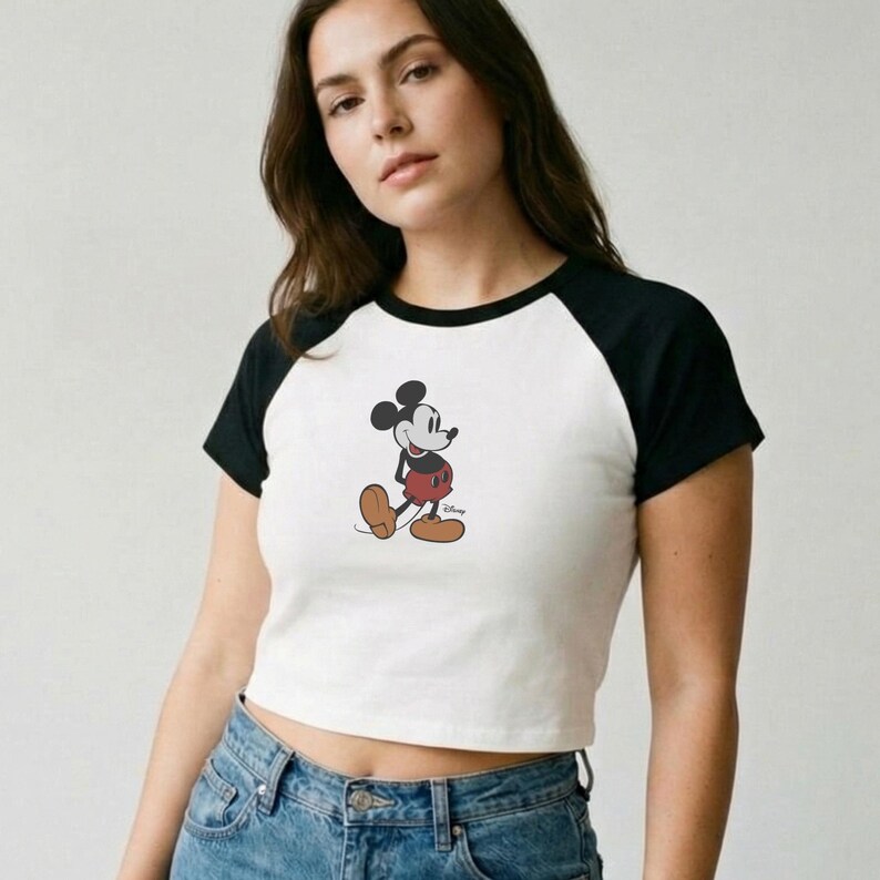 Op de afbeelding: Witte crop top met zwarte raglanmouwen met een Mickey Mouse-afbeelding. Het stripfiguur is afgebeeld in zijn klassieke outfit, met een rode broek, gele schoenen en witte handschoenen. Het Disney-logo is zichtbaar.