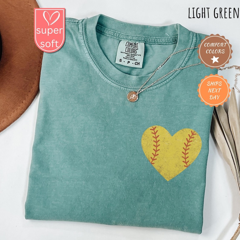 Puede incluir: Camiseta verde claro con un dise&ntilde;o de coraz&oacute;n de softball amarillo. La camiseta tiene el texto "COMFORT COLORS" en la etiqueta. El texto "super soft" est&aacute; en un coraz&oacute;n rosa.
