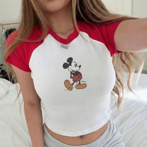 Op de afbeelding: Witte crop top met rode korte mouwen en kraag met een Mickey Mouse-afbeelding. Het Disney-shirt heeft een casual, retro stijl. De top is gemaakt van een zacht materiaal.