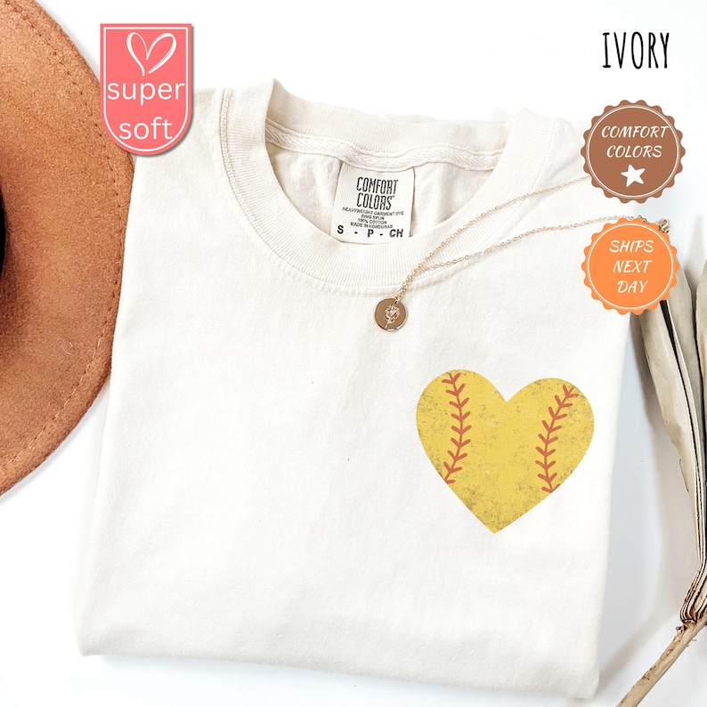 Puede incluir: Camiseta de color marfil con un gr&aacute;fico en forma de coraz&oacute;n de softball. El coraz&oacute;n es amarillo con costuras rojas. La camiseta est&aacute; hecha de una tela suave y c&oacute;moda.