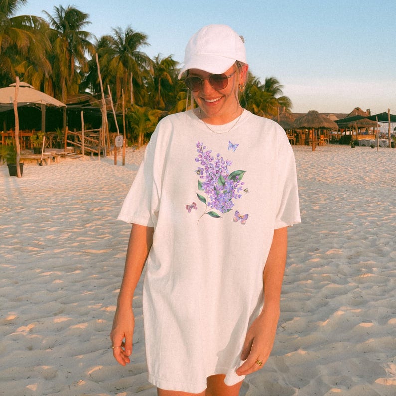 May include: White t-shirt with a purple floral design, butterflies, and a bee. The tee is a relaxed fit, suitable for casual wear. The shirt is made of a soft, comfortable material.