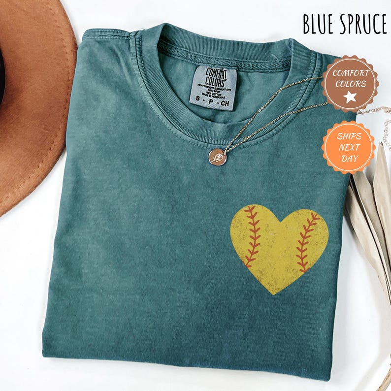 Puede incluir: Una camiseta azul turquesa con un gr&aacute;fico de softball en forma de coraz&oacute;n amarillo. La camiseta tiene un aspecto desgastado y el texto "Comfort Colors" est&aacute; impreso en la etiqueta.