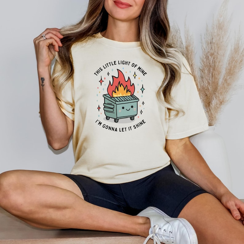 May include: A cream colored t-shirt with a graphic of a green dumpster on fire with a smiling face. The text on the shirt reads "This little light of mine I'm gonna let it shine."