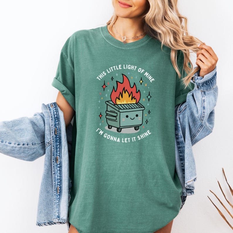 May include: A green t-shirt with a graphic of a cartoon dumpster on fire with the text "This little light of mine I'm gonna let it shine".
