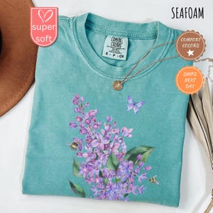 May include: Seafoam green t-shirt with a floral design featuring purple flowers, green leaves, a butterfly, and bees. The shirt has a crew neck and short sleeves. A gold necklace with a pendant is draped on the shirt. The shirt is labelled "Comfort Colors."
