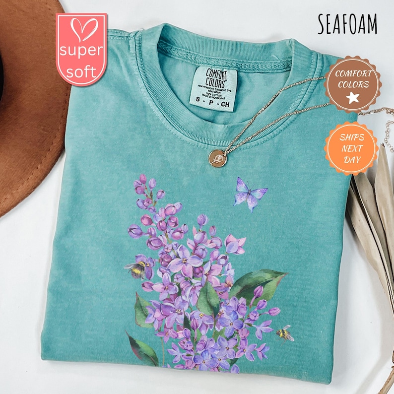 May include: Seafoam green t-shirt with a floral design featuring purple flowers, green leaves, a butterfly, and bees. The shirt has a crew neck and short sleeves. A gold necklace with a pendant is draped on the shirt. The shirt is labelled "Comfort Colors."