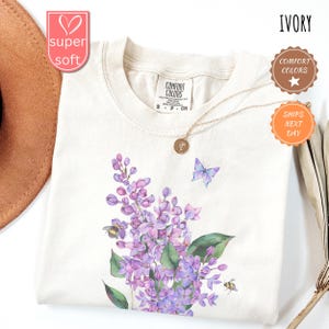 May include: Ivory-coloured t-shirt with a floral design featuring purple lilacs, green leaves, and a blue butterfly. The shirt has a crew neck and short sleeves. A gold necklace with a pendant is draped on the shirt.