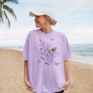 May include: A lilac-coloured t-shirt with a watercolour design of a lilac branch with purple flowers, green leaves, and butterflies. The shirt is made of a soft, comfortable material.