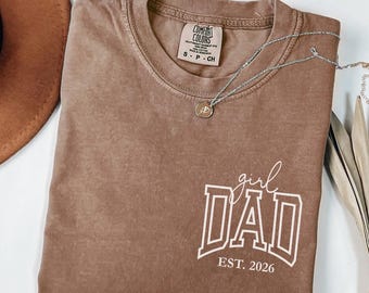 May include: Espresso-colored t-shirt with the text "girl DAD EST. 2026" in white. The shirt has a crew neck and short sleeves. A silver necklace with a pendant is draped across the shirt.