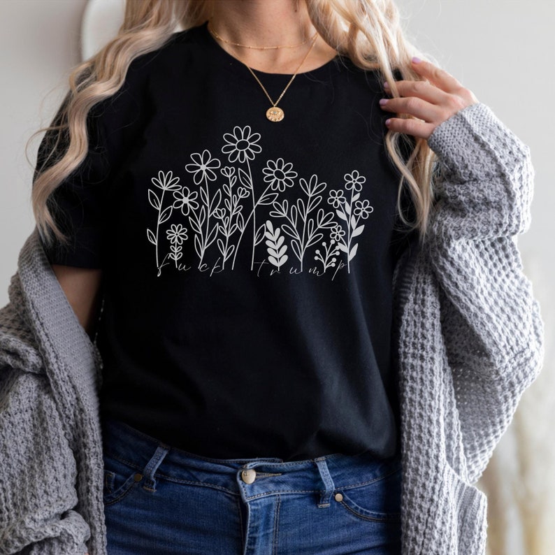 May include: Black t-shirt with a white floral design and the text "F*ck Trump". The graphic features various flower and leaf illustrations. The shirt is made of a soft material.