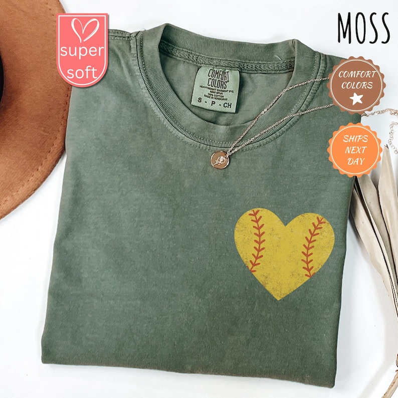 Puede incluir: Una camiseta verde con un gr&aacute;fico de pelota de softball en forma de coraz&oacute;n amarillo. La camiseta tiene un aspecto desgastado y est&aacute; hecha de una tela suave y c&oacute;moda. El texto "COMFORT COLORS" est&aacute; impreso en la etiqueta.