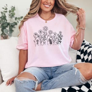 May include: A light pink t-shirt featuring a black floral design with the word "Bloom" at the bottom. The shirt is made of soft material and has a relaxed fit. The design includes various flower and leaf illustrations.