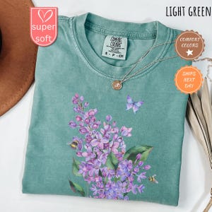 May include: Light green t-shirt with a floral design featuring purple flowers, a butterfly, and bees. The shirt has a crew neck and short sleeves. The text "COMFORT COLORS" is visible on the tag.