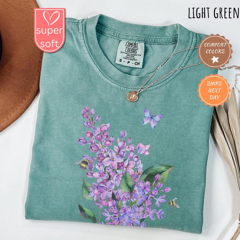 May include: Light green t-shirt with a floral design featuring purple flowers, a butterfly, and bees. The shirt has a crew neck and short sleeves. The text "COMFORT COLORS" is visible on the tag.