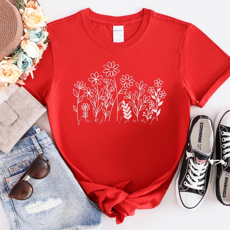 May include: A red t-shirt with a white floral design and the text "F*ck Trump". The shirt is paired with denim shorts, sunglasses, a straw hat, and black Converse sneakers.