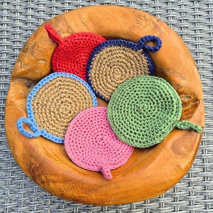 Könnte beinhalten: Eine Holzschale mit fünf handgefertigten Häkeluntersetzern in verschiedenen Farben: Rot, Beige mit blauem Rand, Beige, Grün und Rosa. Jeder Untersetzer hat eine Schlaufe zum Aufhängen.