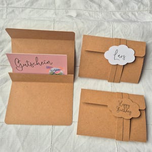 May include: Kraft paper gift card holders with cloud-shaped labels. One holder is open, revealing a pink gift card with the word "Gutschein". Two holders have labels that read "Lars" and "Happy Birthday".