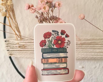 May include: A sticker featuring a stack of vintage books with red and pink flowers growing from the top. The sticker has a light brown background.