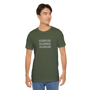 Puede incluir: Camiseta verde oliva con texto de código binario blanco. El texto incluye los números 0 y 1 en una pila vertical. La camiseta está hecha de un material suave y tiene mangas cortas.