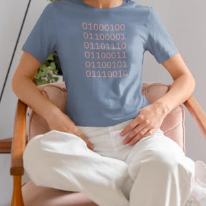 Może przedstawiać: Jasnoniebieska koszulka z różowym nadrukiem kodu binarnego. Kod brzmi: 01000100 01100001 01101110 01100011 01100101 01110010