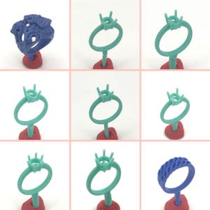 May include: A collection of nine 3D printed wax rings for jewelry making. The rings are in various styles, including a plain band, a ring with a prong setting, and a ring with a detailed design. The rings are all blue or green in color and are displayed on red stands.