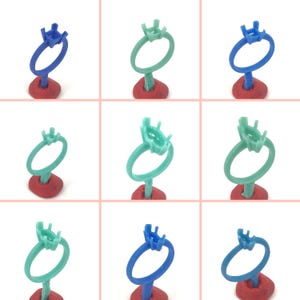 May include: A grid of nine ring molds in various colors, including blue and green. Each ring is mounted on a red base. The rings have a circular band with a square top designed to hold a gemstone. The background is a light pink.