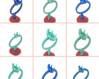 May include: A grid of nine ring molds in various colors, including blue and green. Each ring is mounted on a red base. The rings have a circular band with a square top designed to hold a gemstone. The background is a light pink.