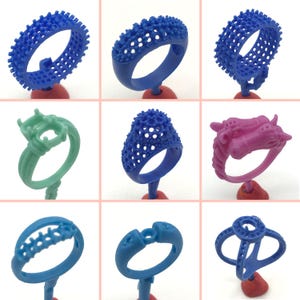 May include: Nine different wax ring models for jewelry making. The rings are in various styles and colors, including blue, green, and pink. The rings are displayed on small stands.