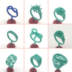 Op de afbeelding: Een verzameling van negen ringontwerpen in verschillende stijlen, gepresenteerd op rode voetstukken. De ringen zijn gemaakt van een wasachtig materiaal in blauw- en groentinten. De ontwerpen omvatten bloemen-, cluster- en geometrische patronen, die sieradenontwerpopties laten zien.