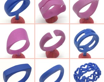 May include: Nine different wax ring molds for jewelry making. The molds are made of a blue or pink wax and are displayed on red bases. The molds are in various shapes, including a simple band, a band with a cut-out, a band with a wavy design, a band with a double line design, and a band with a heart design.