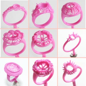 May include: Nine pink wax ring settings for jewelry making. The settings are different styles and sizes, with some featuring intricate designs.