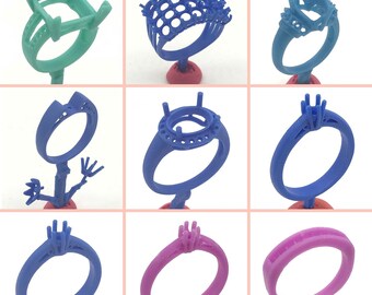 May include: A collection of nine colorful ring molds in various designs. The rings are in shades of green, blue, and pink, each mounted on a small red base. The designs include intricate settings for gemstones, showcasing the variety of ring styles.