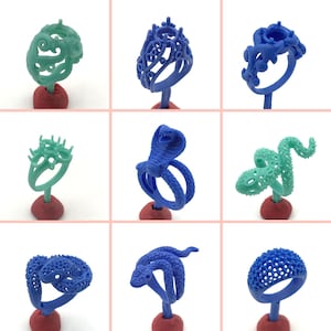 May include: Nine different wax ring designs for jewelry making. The rings are displayed on small blue bases. The rings are made of blue and green wax and feature various designs, including a snake, a crown, and a floral pattern.