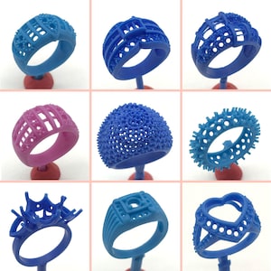 May include: Nine different 3D printed rings in various styles and colors, including blue, pink, and white. The rings are displayed on small stands.