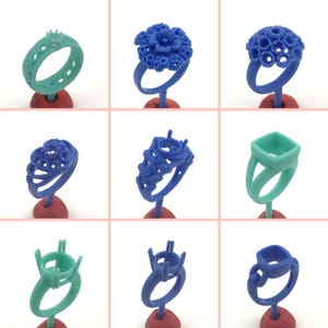 May include: Nine different wax ring designs for jewelry making. The rings are made of blue and green wax and are displayed on small red and green bases.