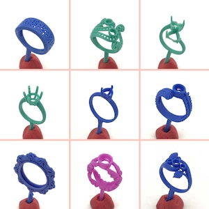 May include: Nine different wax ring designs for jewellery making. The rings are displayed on red bases and are made of blue, green, and purple wax.
