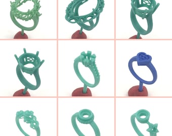 May include: A collection of nine teal and blue ring molds, each mounted on a small red base. The designs vary, including round, oval, and heart-shaped settings, as well as rings with intricate floral and star details. These molds are used in jewelry making.