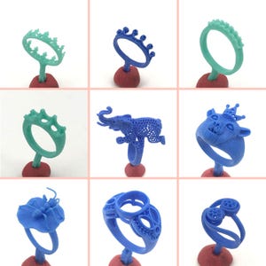 May include: Nine different wax ring designs for jewelry making. The rings are made of blue and green wax and feature various designs, including a crown, an elephant, and a geometric pattern.