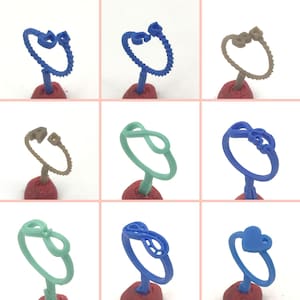 May include: Nine different wax ring designs for jewelry making. The rings are in various colors, including blue, green, and brown. The rings are displayed on small red stands.