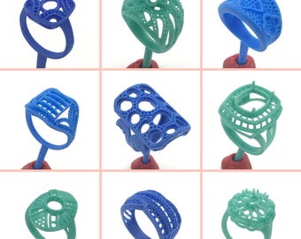 May include: A collection of nine ring molds in various designs and colors. The rings are in shades of blue and green, each mounted on a red base. The designs include openwork patterns, geometric shapes, and textured surfaces, showcasing a variety of styles.