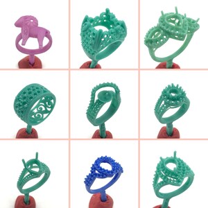 Op de afbeelding: Negen verschillende wasringontwerpen voor het maken van sieraden. De ringen zijn gemaakt van groene was en worden gepresenteerd op rode standaards. De ringen hebben verschillende stijlen, waaronder een kroon, een hart en een eenvoudige band.