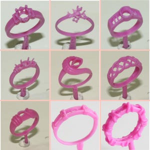 May include: Nine pink wax ring models for jewellery making. The rings are displayed on small white stands. The rings feature different styles and designs, including a ring with a large centre stone, a ring with a snake design, and a ring with a braided design.