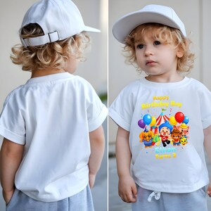 Op de afbeelding: Wit T-shirt met een kleurrijke afbeelding van stripfiguren, ballonnen en een circustent. De tekst "Happy Birthday Boy" en "Ethan Turns 3" staat erop. Het kind draagt een witte baseballpet.