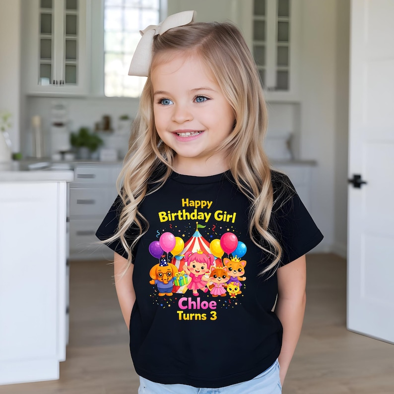 Op de afbeelding: Een zwart t-shirt met de tekst "Happy Birthday Girl" en "Chloe Turns 3" in geel en roze. Het shirt heeft een kleurrijke afbeelding van stripfiguren, ballonnen en een circustent. Het meisje draagt een witte strik in haar haar.