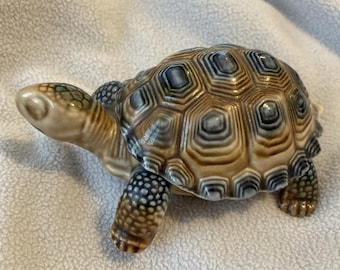 May include: A ceramic turtle figurine with a detailed shell pattern in shades of brown, beige, and blue. The turtle's head and legs are a lighter beige with darker spots. The figurine is a decorative item.