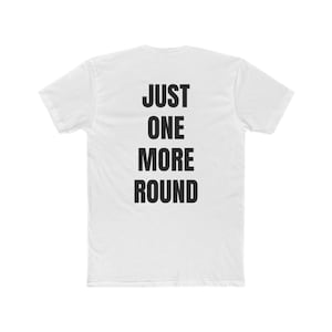 Puede incluir: Camiseta blanca con la frase "JUST ONE MORE ROUND" impresa en letras mayúsculas negras y en negrita. El texto está alineado verticalmente. La camiseta tiene cuello redondo y mangas cortas. Prenda casual.
