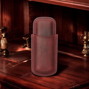 May include: A burgundy leather cigar case with a flap closure. The case is made of high-quality leather and has a smooth finish.