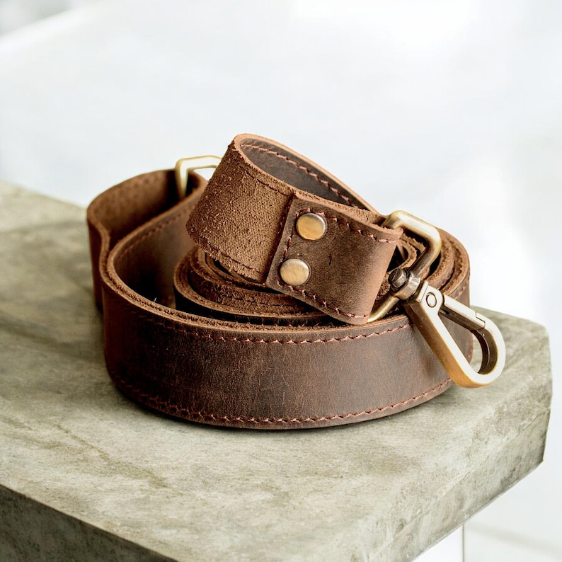 May include: A brown leather camera strap with brass hardware and stitching. The strap is folded and coiled, showing the texture of the leather.
