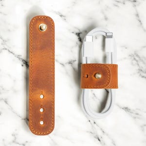 May include: Two brown leather cable organizers with brass hardware. One is a long, rectangular strap with three holes. The other is a small loop securing a white charging cable. The leather has a distressed finish.